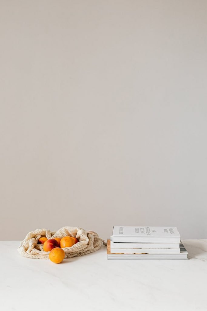 Composition of canvas bag with fresh ripe apricots placed on white table near stack of various books