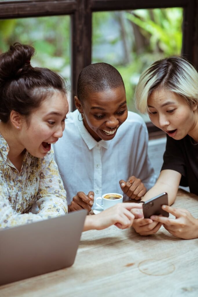 Amazed surprized multiethnic female friends in casual clothes with cup of coffee browsing smartphone while sitting at wooden table with laptop against window